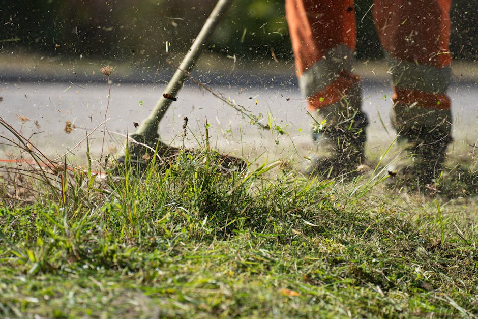 A gardener wearing dark work boots and orange overalls is engaged in lawn maintenance, using a rake or similar tool to clear weeds and debris from a patch of grass. The foreground shows a variety of g