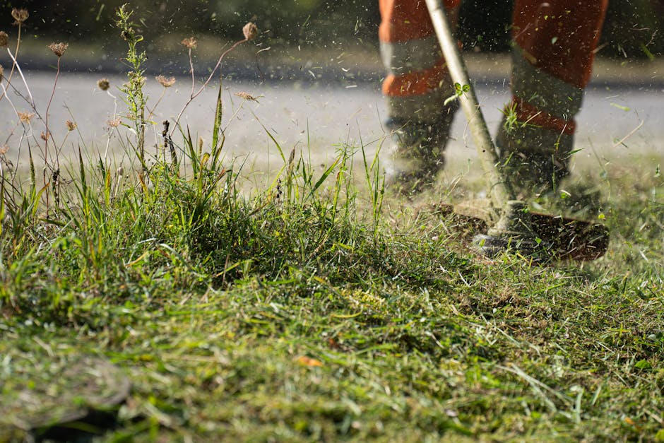 A gardener wearing dark work boots and orange overalls is engaged in lawn maintenance, using a rake or similar tool to clear weeds and debris from a patch of grass. The foreground shows a variety of g