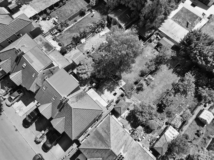 An aerial black-and-white photograph of a residential neighborhood showing closely spaced houses with pitched roofs, driveways with parked cars, and a large communal garden area behind the homes. The