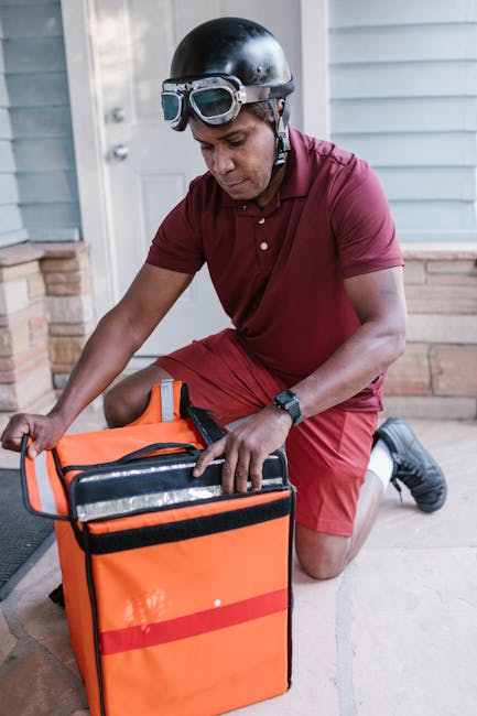 A male delivery worker wearing a black helmet and goggles, dressed in a red uniform, is kneeling on a doorstep as he accesses an insulated orange delivery bag, preparing to deliver a package. The scen